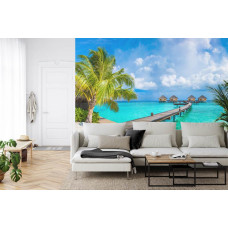 Eine Fototapete mit einer tropischen Urlaubsatmosphäre mit Palmen am Strand und blauem Meer, abgebildet in einem Wohnzimmer.