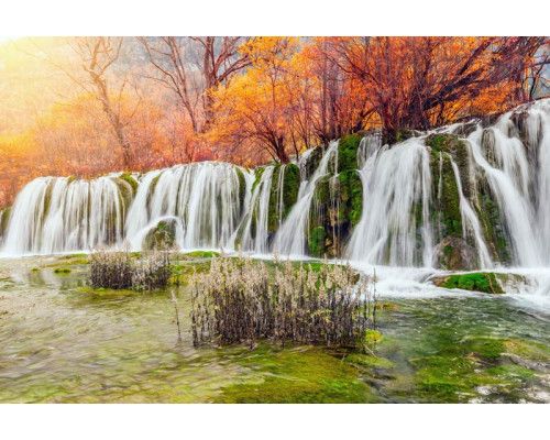 Fototapete mit einem Wasserfall, der durch eine Herbstlandschaft fließt, umgeben von bunten Bäumen und Blättern.
