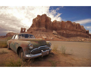 Fototapete eines alten Autos, das in einer Wüstenlandschaft zwischen Felsen steht, mit einem klaren blauen Himmel.
