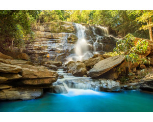 Fototapete eines sonnigen Wasserfalls, der durch Felsen fließt, mit klarem blauen Himmel und strahlendem Sonnenschein.